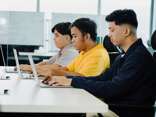 Three young men sit side by side at a desk, working on laptops in a bright office setting with whiteboards in the background.