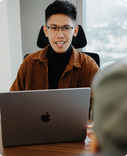 A person wearing glasses and a brown jacket sits at a desk, working on a laptop, with another person partially visible in the foreground.
