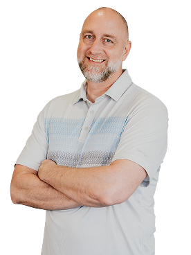 Bald man with a gray beard wearing a light gray polo shirt stands with arms crossed and smiles at the camera against a plain white background.