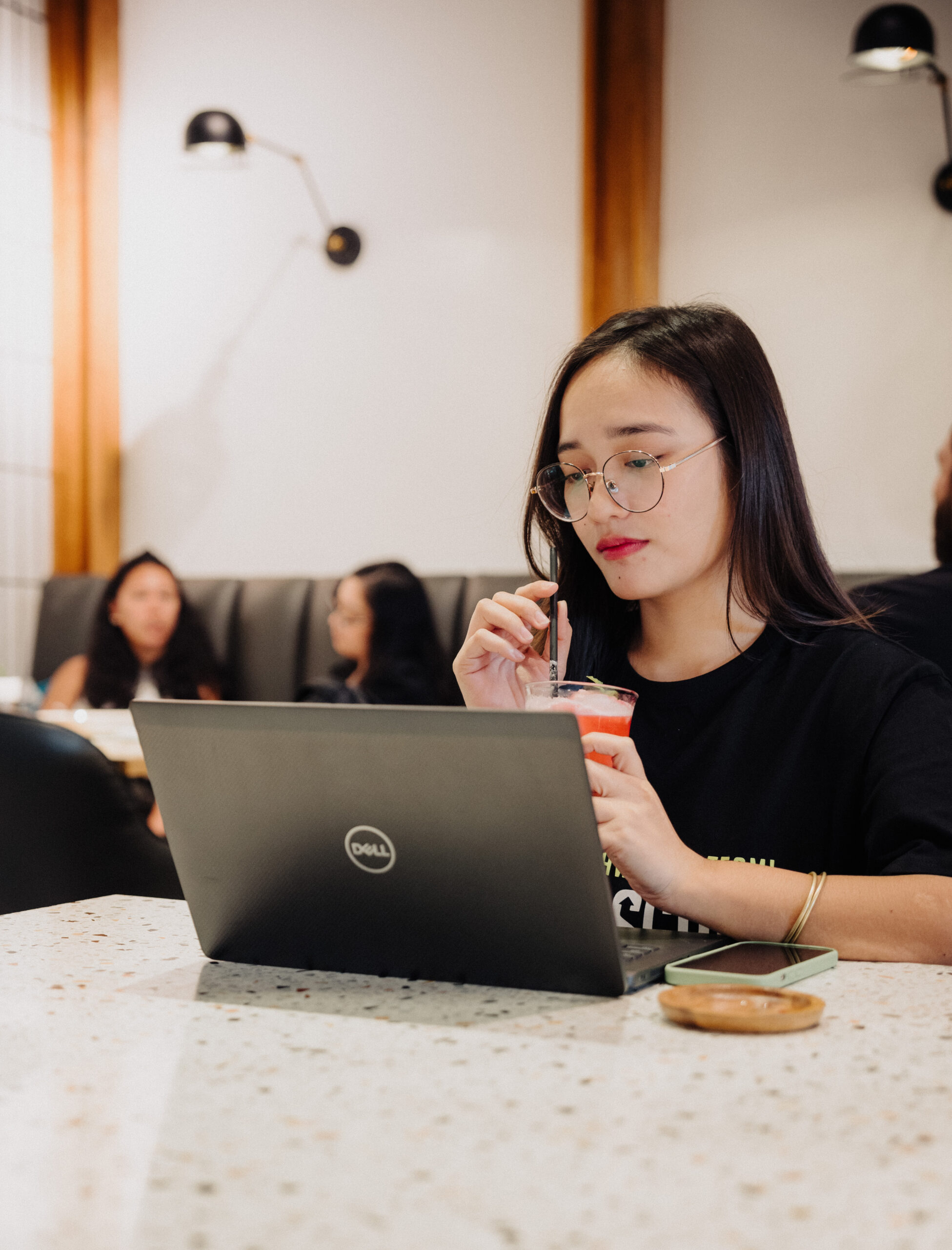 A woman with glasses works on a Dell laptop at a café, sipping a drink through a straw. Other people are seated and talking in the background.