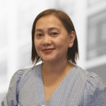 A woman with straight, shoulder-length hair and a light blue checkered blouse smiles slightly in front of a blurred background, embodying the confidence often seen in product operations professionals.