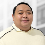 A man with short black hair wearing a cream-colored collared shirt stands in front of a blurred, light-colored background, representing expertise in custom software development services.