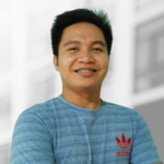 A man with short black hair, wearing a blue Adidas t-shirt, stands smiling with arms crossed against a blurred indoor background, reflecting the confidence and professionalism seen in top-tier software testing services.