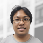A person with short black hair and glasses, wearing a light gray shirt, stands in front of a blurred background with large windows—representing skilled .net developers for hire ready to deliver top-quality solutions.