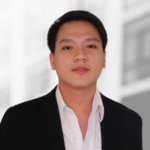 A young man in a black blazer and white shirt faces the camera with a neutral expression; the background is blurred.