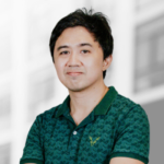 A person with short black hair wearing a green patterned polo shirt stands in front of a blurred building background.