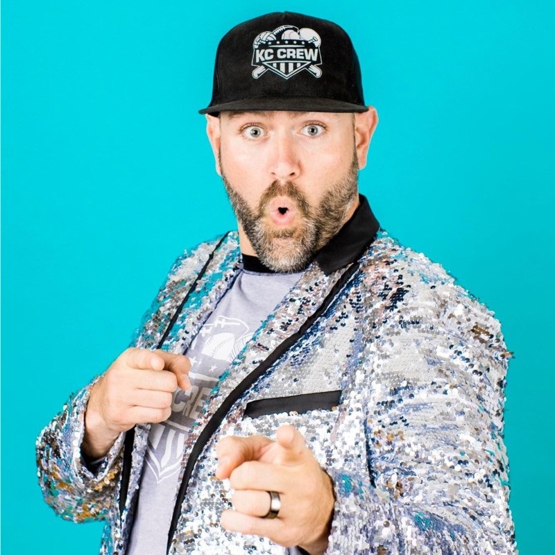A man in a sequined jacket and KC Crew hat points at the camera with an open-mouthed, surprised expression in front of a bright blue background. A man in a sequined jacket and KC Crew hat points at the camera with an open-mouthed, surprised expression in front of a bright blue background.