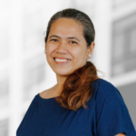 A woman with long brown hair tied back, wearing a navy blue top, smiles at the camera with a blurred building in the background.