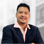 A man in a dark blazer and light pink shirt smiles at the camera with a blurred building background, reflecting confidence often seen in leaders of SaaS platforms.