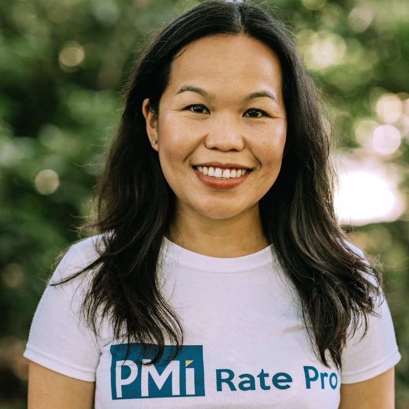 Smiling woman with long dark hair wearing a white "PMI Rate Pro" t-shirt stands outdoors in front of greenery. Smiling woman with long dark hair wearing a white "PMI Rate Pro" t-shirt stands outdoors in front of greenery.