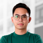 A young man with short dark hair, wearing round eyeglasses and a green shirt, stands in front of a blurred building—ideal for those looking to outsource mobile app development or hire mobile app developers for their next project.